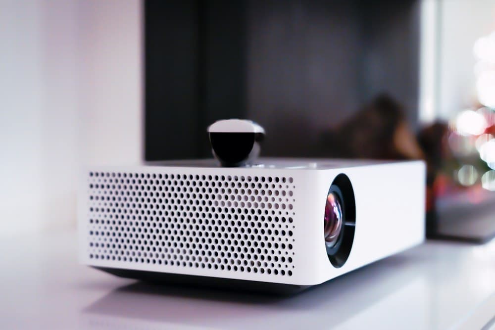 Compact white home cinema projector on a shelf, lens towards the camera